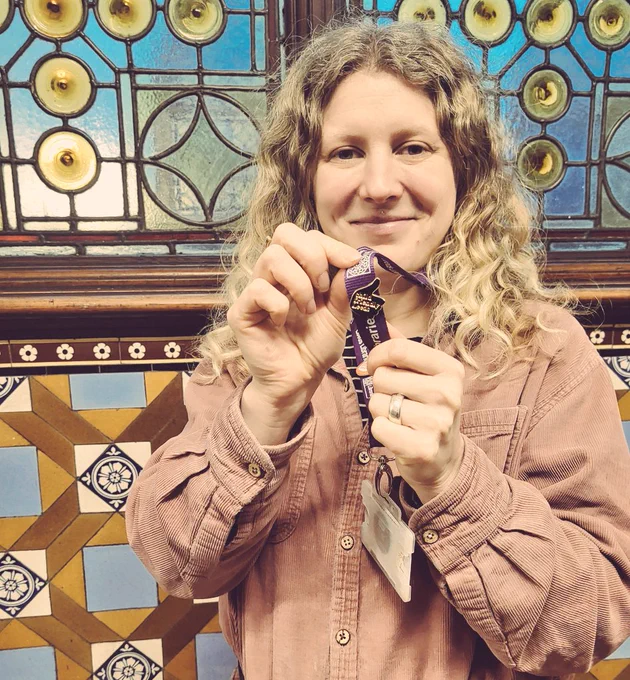 Rachel stands in front of a stained glass window, smiling to camera holding her gold thumbs up badge from Child Friendly Leeds
