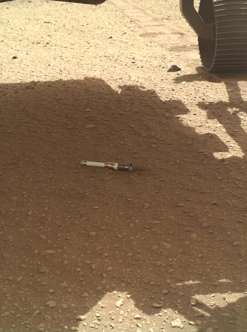 Photo from NASA’s Mars Perseverance rover shows a small, white sample tube lying on the sandy surface of Mars. About the size of a marker, the tube sits in the shadow of the rover that released it. Part of the rover’s left rear wheel is visible at top right, along with tracks it has made on the dusty ground.