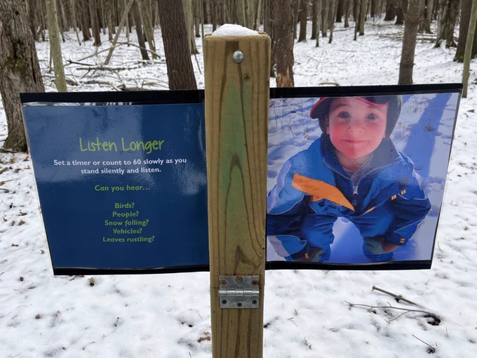 Sign along trail with a picture of a child playing in the snow and a caption saying “Listen Longer. Set a timer or count to 60 slowly as you stand silently and listen.
Can you hear..
Birds?
People?
Snow falling?
Vehicles?
Leaves rustling?”