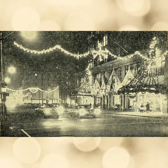 A 1960s black and white newspaper photo of Malley's and Chapel Street with Christmas lights on the buildings and lights strung across the streets in the air. The lights are very visible in the night sky. Two 1960s cars sit at the intersection with their headlights facing the camera.