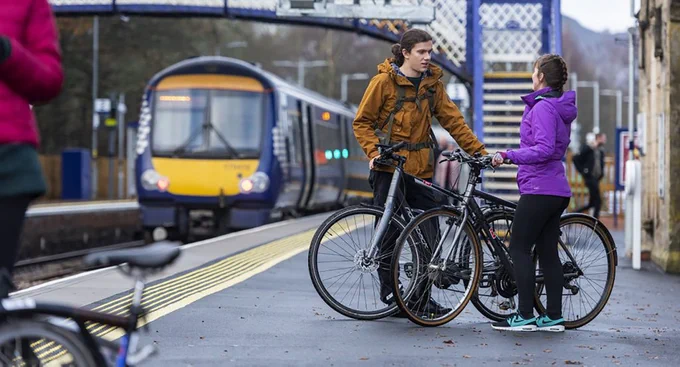 Two people standing with bikes on a railway station platform whilst a ScotRail train arrives