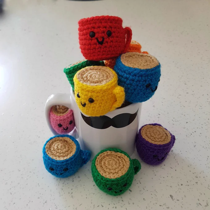 A large coffee mug filled with lots of small crocheted coffee mugs. They all have embroidered faces: some smiling and some grumpy
