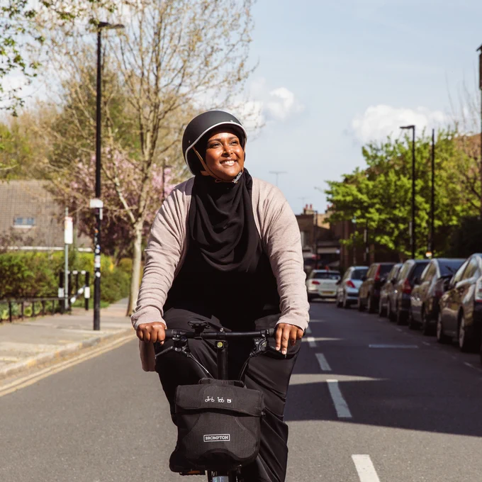 A woman riding her bike and smiling. The sun is shining and there is blossom in the trees in the background