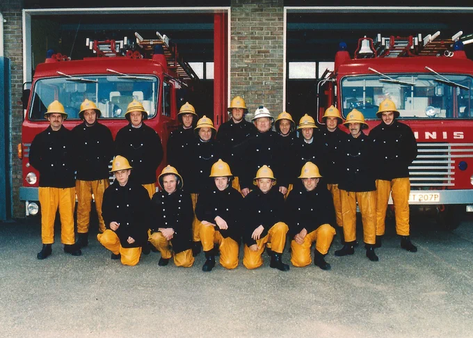17 white men, aged 20 to 60, in firefighter uniform of yellow trousers and navy jackets and yellow firefighting hats, standing in front of two red fire engines parked in a fire station.