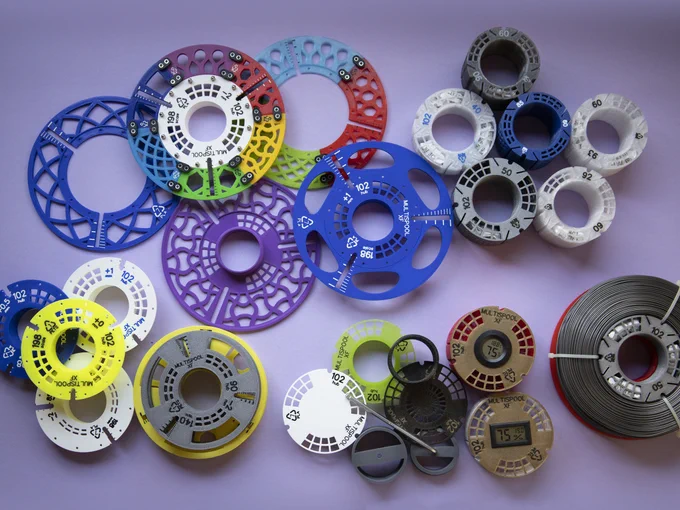 Overall view of a heap of components of the MultiSpool XF spooling system. There are 5 plates or plate disks on top left of the image; 6 cylindrical hubs, with an inner nut-like screw, top right; plate caps and two smaller spools of 140 and 160mm at the bottom left; 7 accessories for hubs at the bottom mid-right; and a filament coil in a partially mounted spool at the bottom far right, not fully visible in the image.