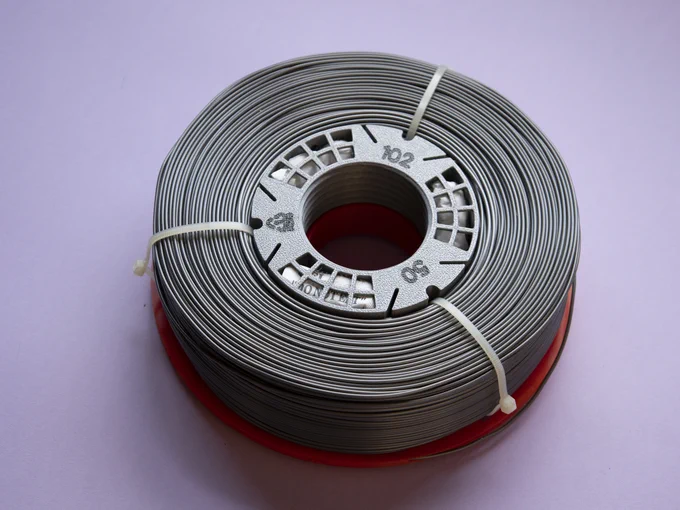 Filament coil in a partially mounted MultiSpool XF spool, with only the bottom plate and the hub; the top plate is missing. Coil has still non-reusable zipties tied. The hub has visible numbers: 102 for the hub diameter, 50 for the hub height; plus there is a PETG recycling symbol with code 1. The hub has 3 120-degree-wide grills, for the desiccant pocket bottoms, and white bags of desiccant can be seen inside the pockets. The filament coil end tip is plugged in a channel in the bottom spool plate.