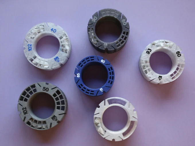 Six different MultiSpool XF core hubs. Five show their bottom, with numbers and symbols on it. Hub dimensions, as shown by their numbers, are: 102mm diameter, 40mm tall; 102mm diameter, 50mm tall; 93mm diameter, 60mm tall; 90mm diameter, 60mm tall; 85mm diameter, 60mm tall. All those show a recycling symbol for PETG with code 1. Last hub is viewed from top, so its dimensions are unknown (although it's actually 92mm diameter, 60mm tall), and the three desiccant pockets are clearly seen; one has a desiccant bag in it.