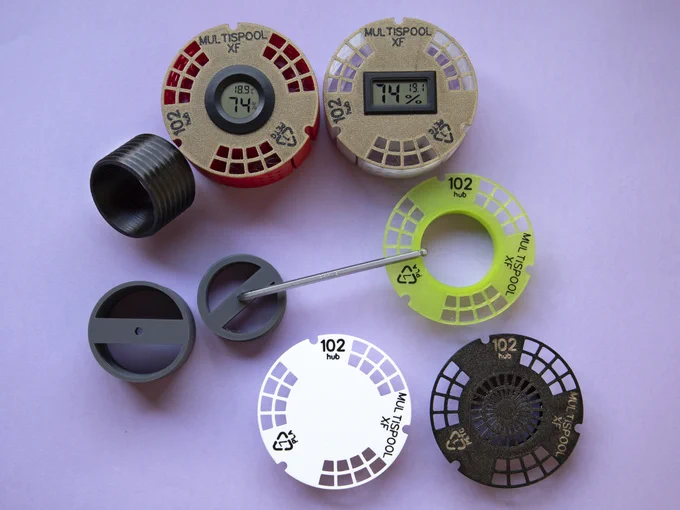 View of the hub accessories. On top of the image there are two hub caps mounted on hubs; they both have a hygrometer (one hygrometer is round, the other rectangular). Below them, left, there is a hub connector: a simple holed screw. Below the hub connector there are two reamers, one for plate caps, the other for hubs. The hub reamer, with a bolt-like screw, has an Allen key inserted in it, to help with turning. At the mid-left and bottom left of the image there are three other hub caps: one has a hole in it, the plain cap, which just closes the desiccant pockets; bottom middle there is a full hub cap, with the hole closed, and bottom right there is a desiccant cap, with a radial grill.