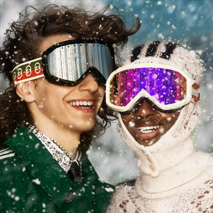 A close up of a Black woman wearing a wool white hood and a white ski mask with GG monogram reflective lenses next to the White man with long curly hair wearing a black ski mask with a red and green striped band with Interlocking G details. Both are smiling as snow falls.  