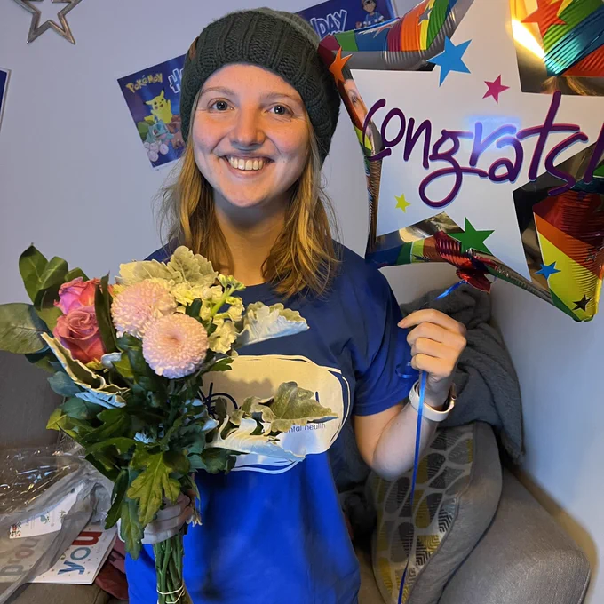 Charlie is smiling and holding flowers and a balloon after doing 24 half marathons