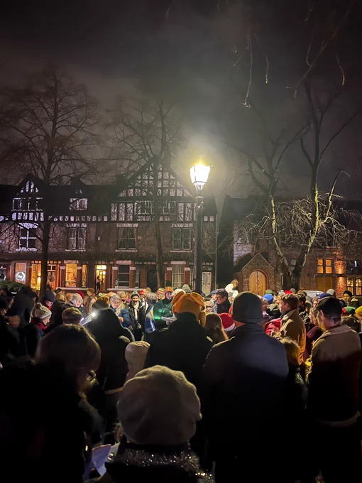 Carol singers gather around a lamp on a green in a suburban area at night. Their heads lifted in song.