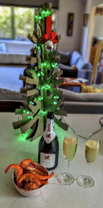 A bowl of cooked prawns and a bottle with two glasses of 2015 Seppelts Salinger sit in front of a small wooden XMas tree with some green fairy lights and other decorations on it.