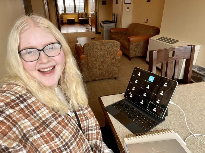 ACBS President Olivia O’Connell sits at a table with her iPad open to a Zoom call. She is smiling at the camera. Olivia is wearing a brown, square patterned shawl over a white shirt.