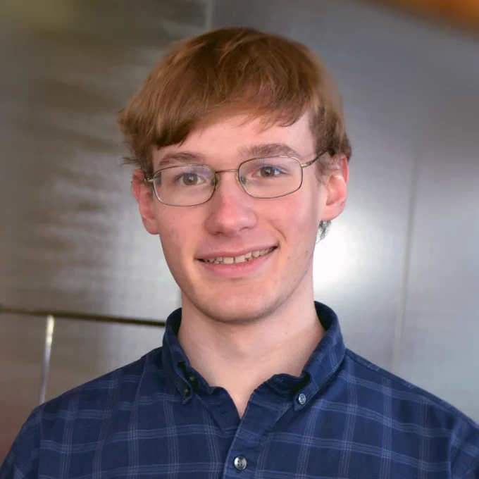 A photo of Thomas from the shoulders up in front of an unidentifiable background (probably a wall? it's brownish but reflective?). He's smiling at the camera and wearing glasses and a blue plaid(?) collared shirt.