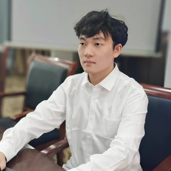 A photo of Zhewen seated at a table (desk?). He's wearing a white collared shirt and looking away from the camera with a neutral/focused expression, probably towards the front of the room. The table appears to be made of a dark wood, and the chair (4 legs, 2 arms) matches the table, with a square of dark leather(?) padding on the back of the seat. There's another chair next to Zhewen at the table in the background of the photo, but it's empty.