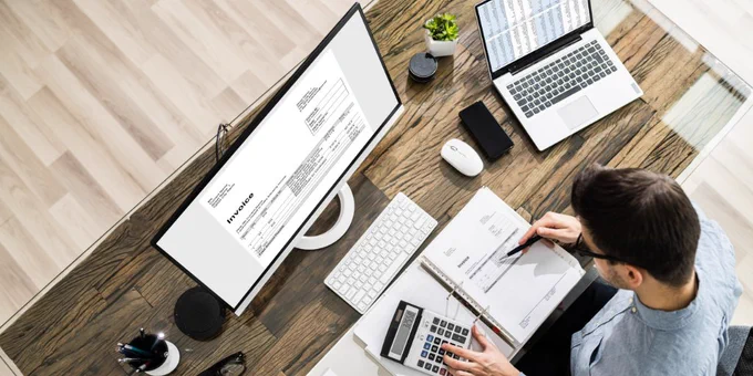 A man sits at a desk with 2 monitors. One has an invoice, and the other has spreadsheets.