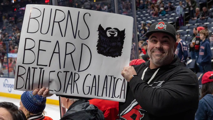 Photo of a fan at the game holding up a sign that says, "Burns. Beard. Battlestar Galatica."