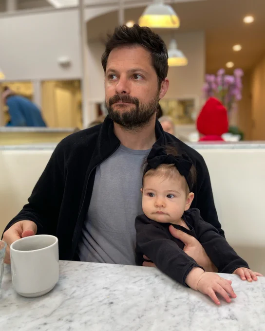 Doug holds his baby girl as the both look off to the left. Doug holds a cup of coffee in his other hand. They both wear black and a seated in a brightly lit diner