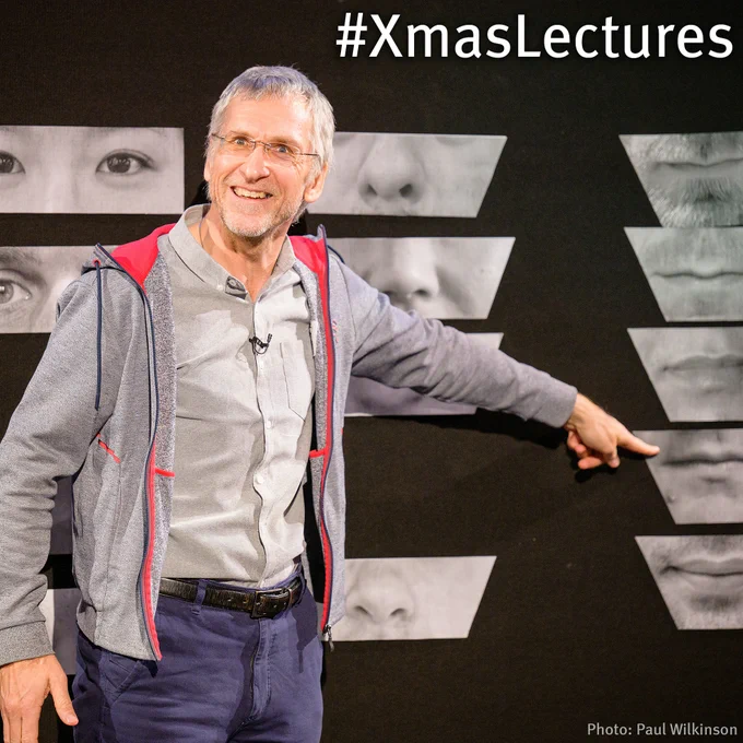 Charlie Frowd points at a wall of images of facial features. He is smiling and wearing a grey hoodie and shirt.