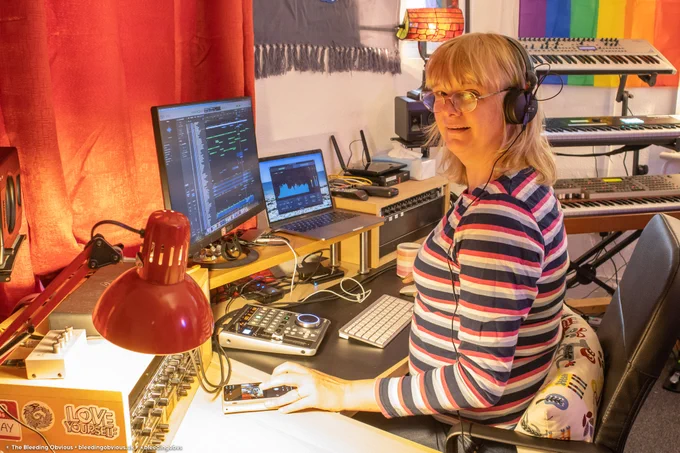 A woman sitting in a recording studio, wearing headphones and using a computer. In the background are synthesisers. She is smiling at the camera.