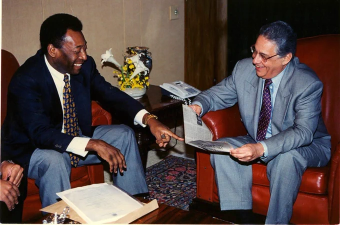 Fotografia colorida que mostra Fernando Henrique Cardoso e Pelé. Eles estão sentados; um de frente para o outro; sorriem. À esquerda, Pelé, que veste terno, calça social e camisa branca. Com a mão esquerda, ele aponta para um recorte de jornal que está nas mãos de Fernando Henrique Cardoso, que veste terno, calça social e camisa; FHC usa óculos. Ao fundo, uma pequena mesa com um vaso de flores e um telefone com fio. 