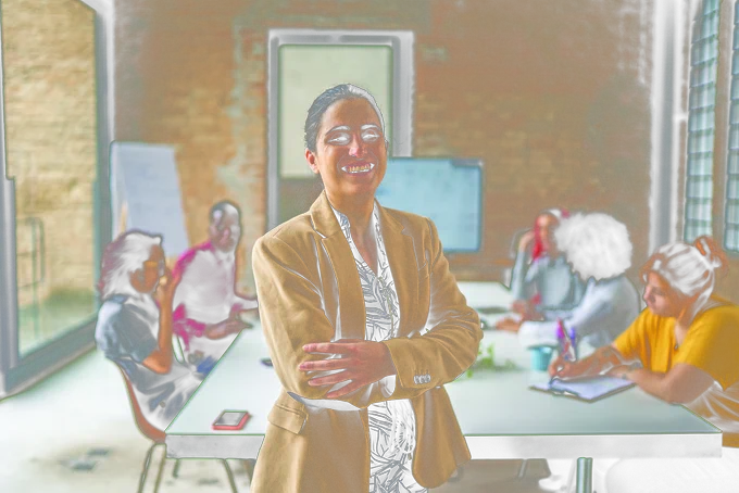 A young professional person who is blind stands in the front of a board room. She smiles with her arms crossed proudly. 