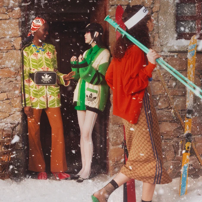A white woman with long wavy brown hair under a white Adidas headband, carrying skis and poles, walks under falling snow past a Black woman with a GG Trefoil silk scarf and an Asian woman with a black riding helmet, both chatting in the doorway of a stone chalet. They are all wearing adidas x Gucci looks including a long GG pattern skirt with blue stripes, a sweater dress and belt bag with the Gucci Trefoil, and a felted wool jacket with graphic wave and GG Trefoil pattern.  