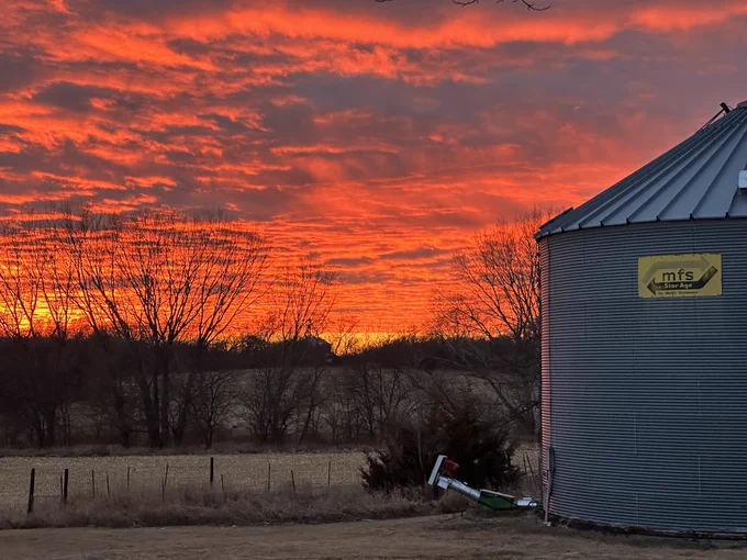 A blazing orange sunset. The sky is filled with clouds. In the foreground, a silver grain silo sits at the right side of the photo, In the background, the silhouettes of leafless trees border rolling fields of harvested corn.