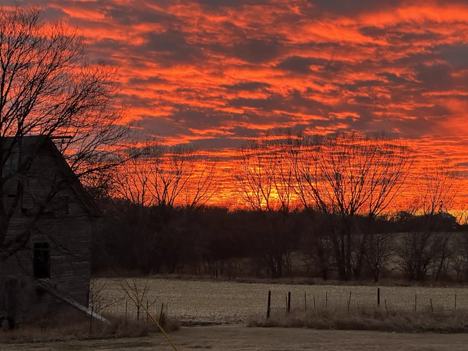 A blazing orange sunset. The sky is filled with clouds. In the foreground, a barn sits at the left side of the photo, its silhouette dimly-lit. Behind it, leafless trees and rolling fields of harvested corn.