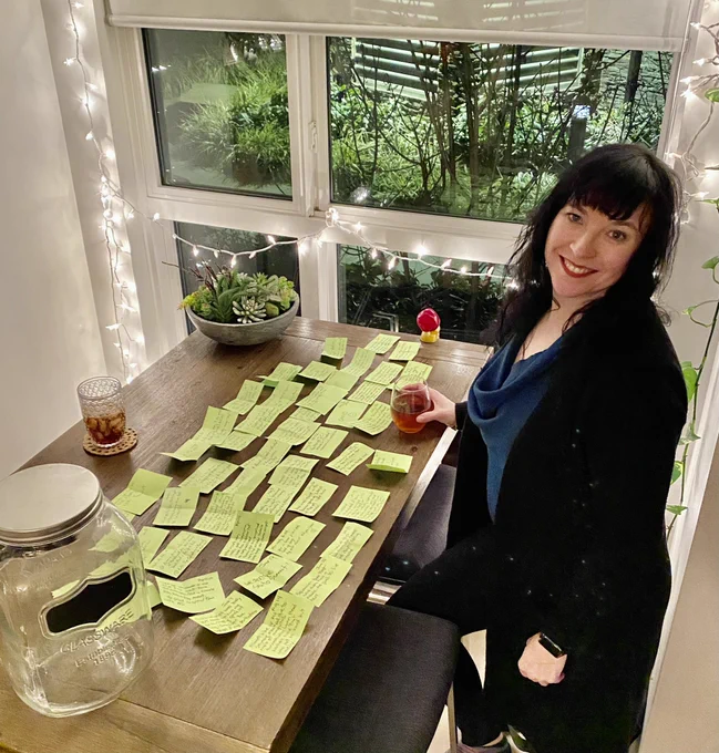 Trish stands at dining table, which is mostly covered by small paper notes laid out chronologically