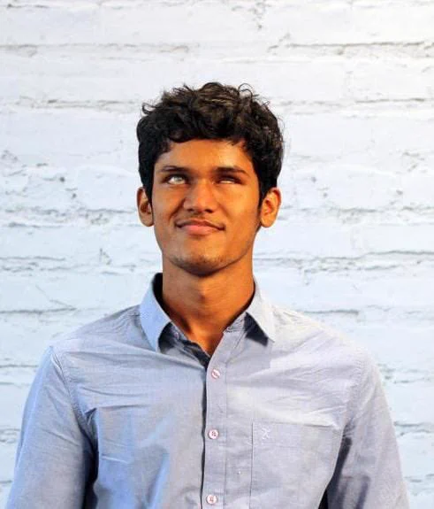 This photo shows Bhavya standing in front of a white brick wall. He is wearing a pale blue button down shirt and looking at the camera.