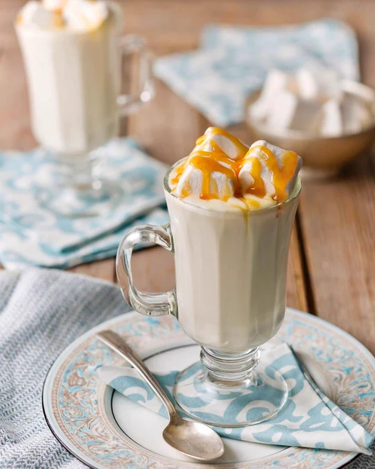 Atop a napkin on a plate sits a glass mug of White Hot Chocolate topped with marshmallows and caramel.