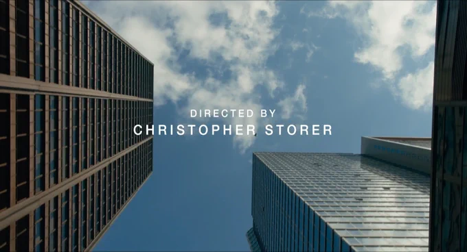 A still from the opening title sequence of The Bear Season 1 Episode 7 showing a view looking up at blue sky and white clouds between tall skyscrapers, the credit for DIRECTED BY CHRISTOPHER STORER in sans serif white text on top.