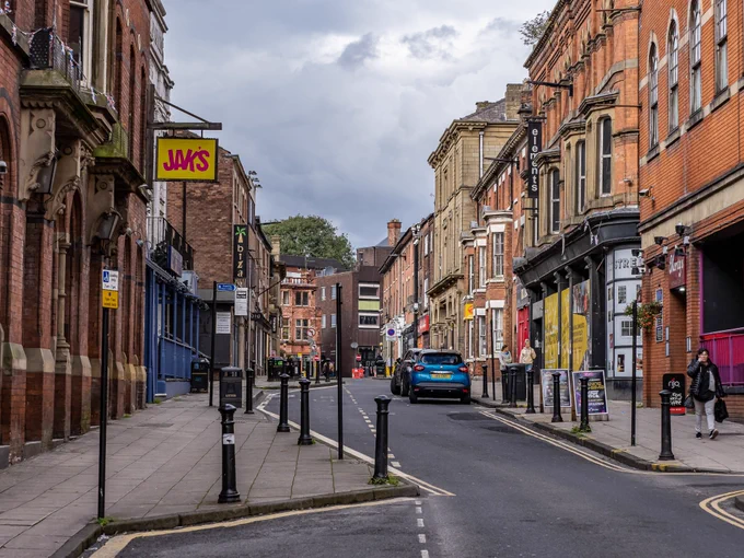 Looking down the middle of King Street in Wigan. Brick buildings on either side of the road with wide pavements. Bollards line the edge of the pavement and people are pictured walking down the street, toward the camera. There is a blue car parked halfway up the street. The sky is cloudy.