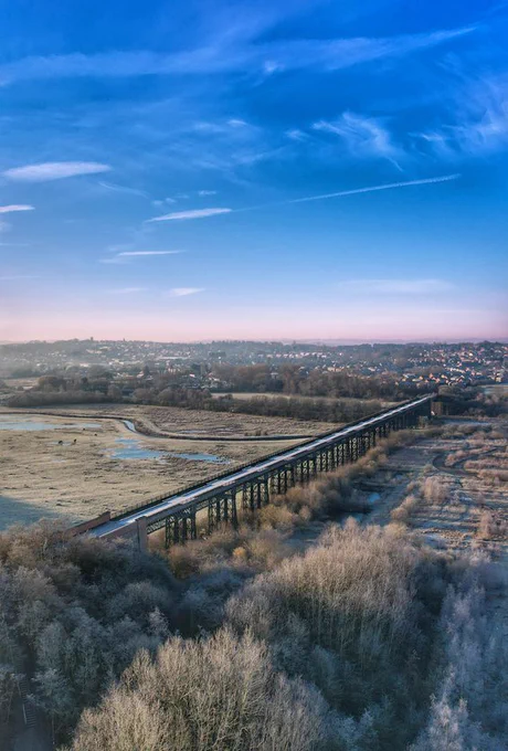 A winter image taken from a drone high above Bennerley Viaduct. The sky is vivid blue with wispy clouds, the fields and trees are brown. The viaduct crosses diagonally from bottom left towards middle right, a straight line in the landscape, with the lattice piers visible beneath. In the distance you can just make out the buildings of Ilkeston. 
