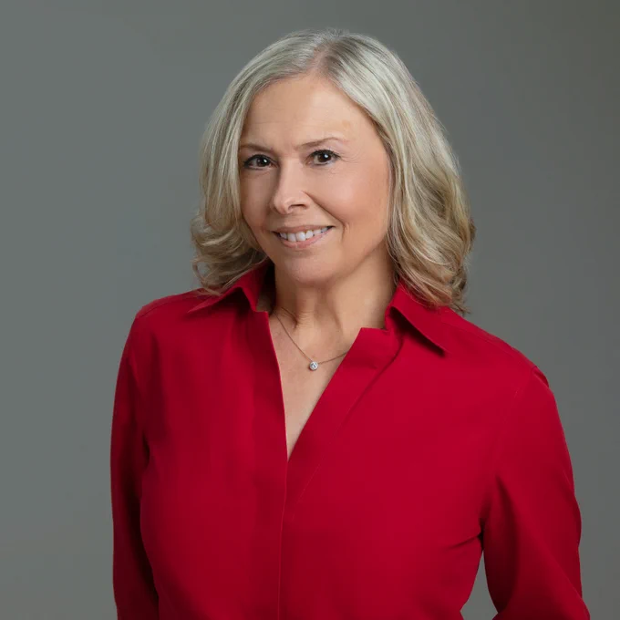 A portrait of Jan Eberle.  Jan is smiling and wearing a red long sleeve top, her blonde hair is a medium length. Around her neck is a diamond pendant on a thin gold chain.