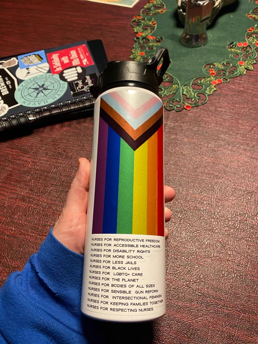 A white water bottle with a vertically aligned progress pride flag, with the triangle of black brown and trans colors set over the rainbow. Below it is a white sticker which reads a laundry list of “nurses for ______” (various progressive causes the character limit for this description won’t allow)

