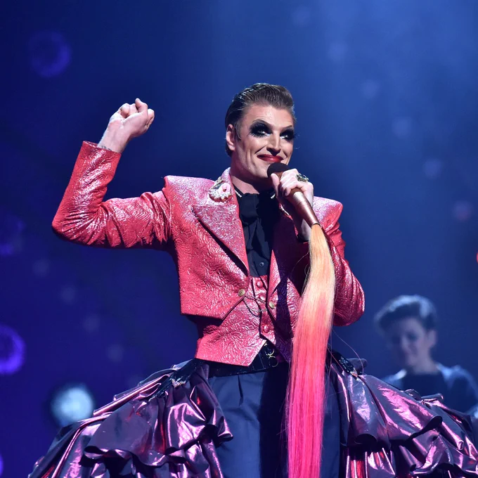 Reuben Kaye performing on stage holding a microphone. They are wearing a pink glittery blazer. 
