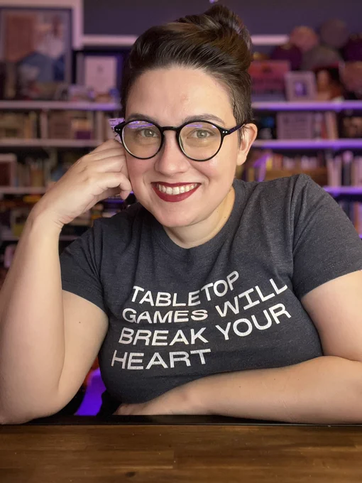 Emily Friedman smiles a camera. She is sitting at her desk wearing dark room to blue light glasses. Her gray blue shirt reads “tabletop games will break your heart”