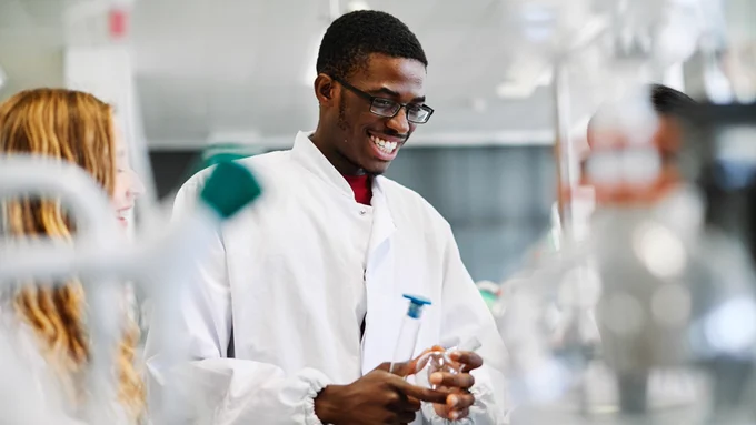 Two students are in a lab, possibly in the middle of an experiment. The person in the center holds a large volumetric flask filled with blue liquid. In front of them and out of focus are other flasks and lab equipment. 