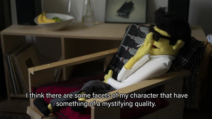 A yellow puppet wearing sunglasses and a white turtleneck reclines on a chair whilst smoking a cigarette. White text overlays the image in the style of subtitles and states: "I think there are some facets of my character that have something of a mystifying quality".

The images is a production still from Joshua's film ‘Pier Paolo (a portrait)’ from 2021.