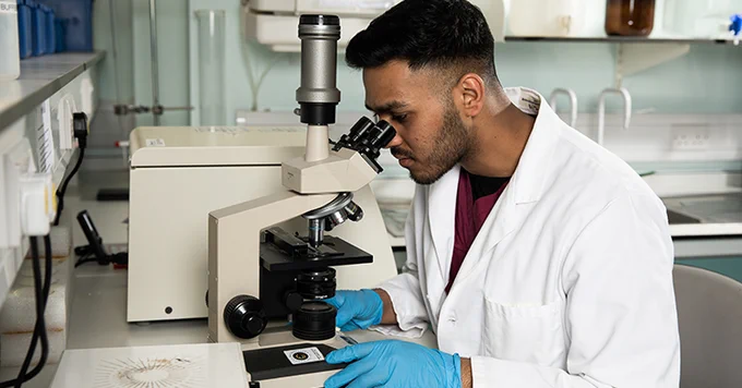 PhD student wears a lab coat and looks through industrial microscope.