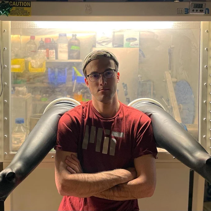 A photo of Ashton standing arms crossed and unsmiling in front of a glove box. The glove box appears to be lit, but the room itself appears poorly lit, and the resulting lighting gives the photo a dramatic feel. Ashton is wearing a red short-sleeve MIT t-shirt and a backwards baseball cap. He's also wearing glasses, but not safety glasses, which raises the question of why he's in a lab (not ours!) without at least proper eye protection on.