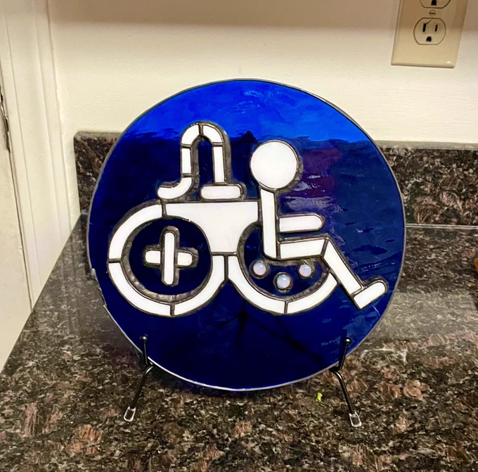 Stained glass piece displayed in a stand on a kitchen countertop of the game accessibility joy rider logo inside of a deep blue circle background.