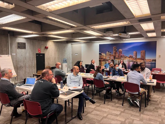 City of Boston DoIT employees are sitting in a conference room during their ITIL-4 training. There is a picture of the city of Boston Harbor Skyline behind them. There are desks with training materials.