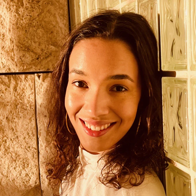 A photo of Collette from the shoulders-up, which looks like it was professionally done. She's smiling at the camera with her head turned a little over her shoulder, and the lighting is a lovely golden color. She appears to be standing in front of a corner where a stone brick (?) and glass wall meet, and she's wearing a white turtleneck. 