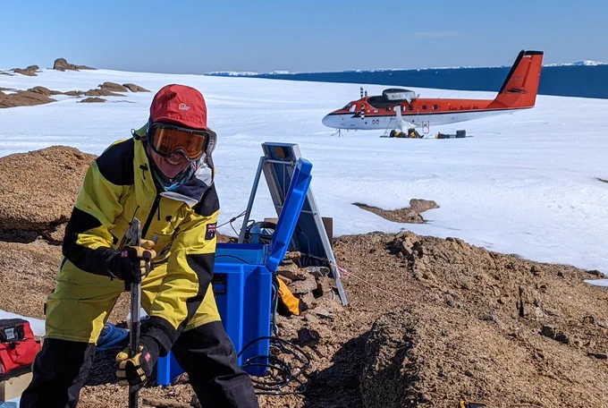 Geophysicist using crowbar to install seismic station on rock. Snow and twin-otter aircraft in background.