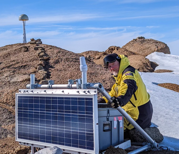 GNSS station on rock showing antenna, snow, solar panel and geophysicist in cold-weather clothing.