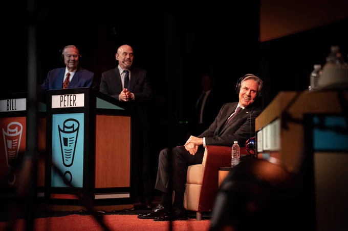 Secretary Blinken is sitting, looking to his left, and smiling. In the background are Peter Sagal and Bill Kurtis standing at podiums.