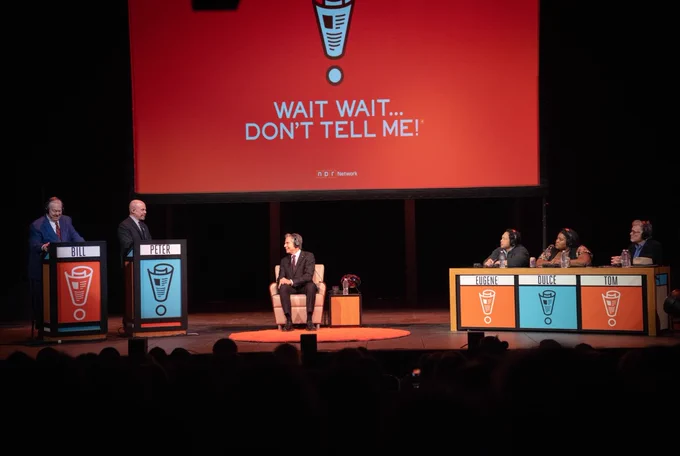 Secretary Blinken is sitting in the middle of a stage. To his right are two podiums with the names “Peter” and “Bill” with Peter Sagal and Bill Kurtis standing at those podiums. To the Secretary's left are three contestants. 